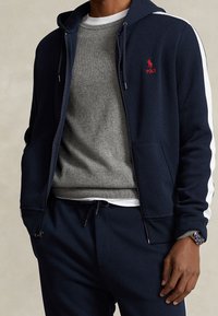 Marinblå zip-up hoodie med vita sidränder, bärs över en grå tröja, ihop med marinblå sweatpants och en klocka. Slät textur, avslappnad design.