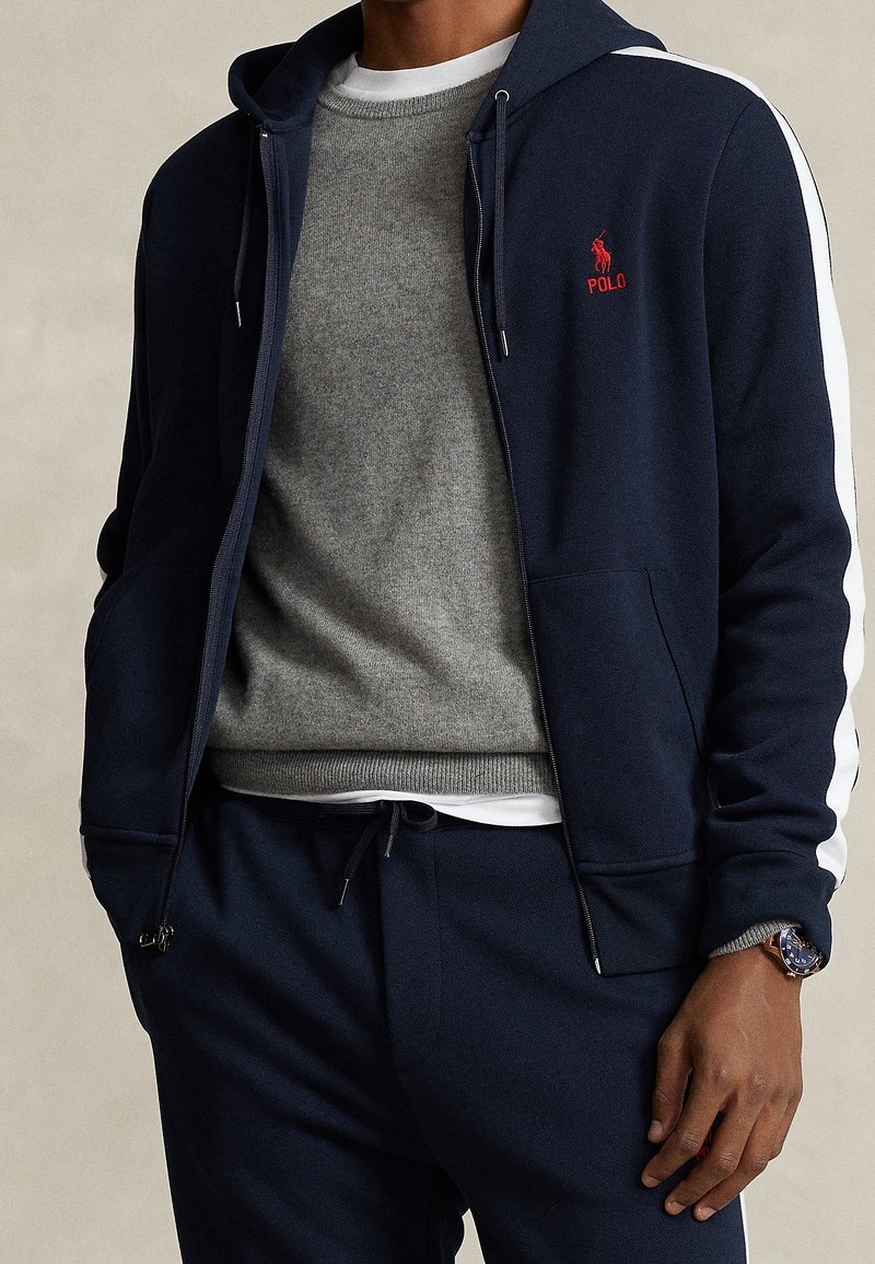 Marinblå zip-up hoodie med vita sidränder, bärs över en grå tröja, ihop med marinblå sweatpants och en klocka. Slät textur, avslappnad design.