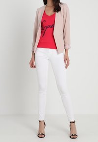 Lyserød blazer over en rød ærmeløs top med sort glitrende tekst, parret med hvide skinny jeans og sorte stilethæle med nitte-accenter.