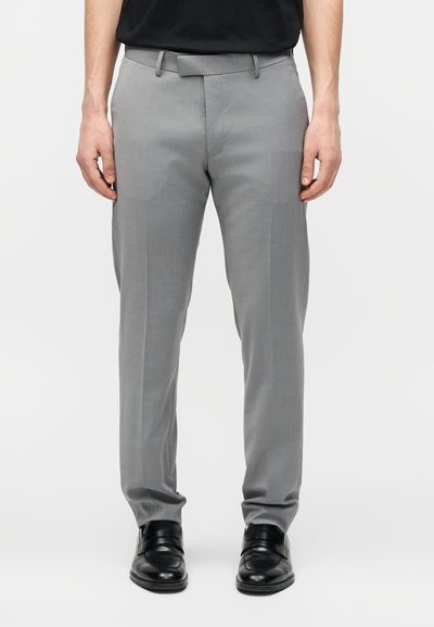 Άνδρας που φοράει ανοιχτό γκρι παντελόνι με πιέτα μπροστά, συνδυασμένο με μαύρα δερμάτινα παπούτσια και μαύρο πουκάμισο σε λευκό φόντο.