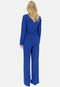 Niebieski garnitur szyty na miarę z dopasowaną marynarką i szerokimi spodniami. Gładka tkanina, strukturalny design i czyste linie. Widoczne szwy i detal z tyłu.