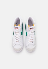 Vita högdesigner sneakers med gröna accenter, gula detaljer och texturerad grå mocka vid tån. Har snörning och en framträdande Nike-logga.