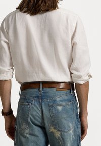 Lehká bílá lněná košile s vyhrnutými rukávy, doplněná o potrhané modré džíny s viditelným šitím a hnědým koženým pásem.