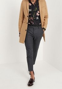 Beige jas over een bloemen zwart shirt, grijze op maat gemaakte broek en donkere schoenen. Kenmerken zijn onder andere een notch lapel en patroon details.