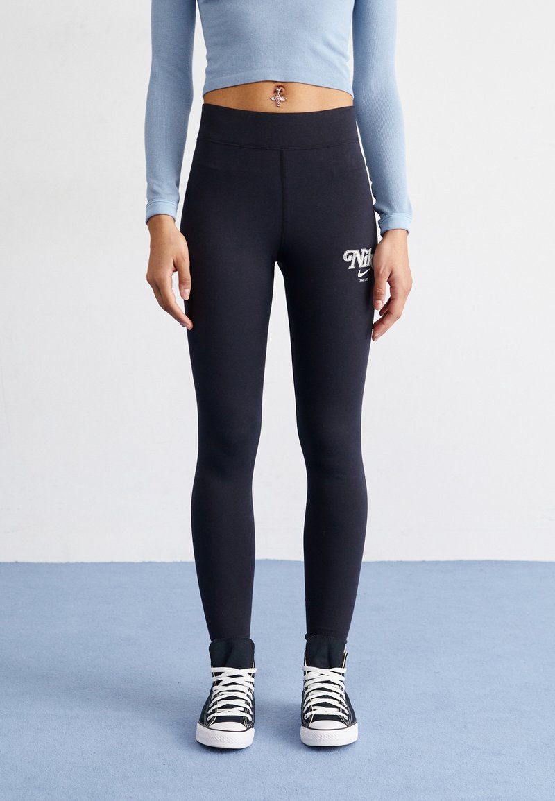 Une personne portant des legging Nike noirs et des baskets montantes noires avec des lacets blancs, se tenant sur un sol bleu contre un mur blanc.