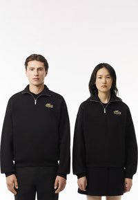 Sudaderas con cremallera negra de material suave, con un logo verde de cocodrilo de Lacoste en el lado izquierdo del pecho. Los cortes del modelo varían ligeramente.