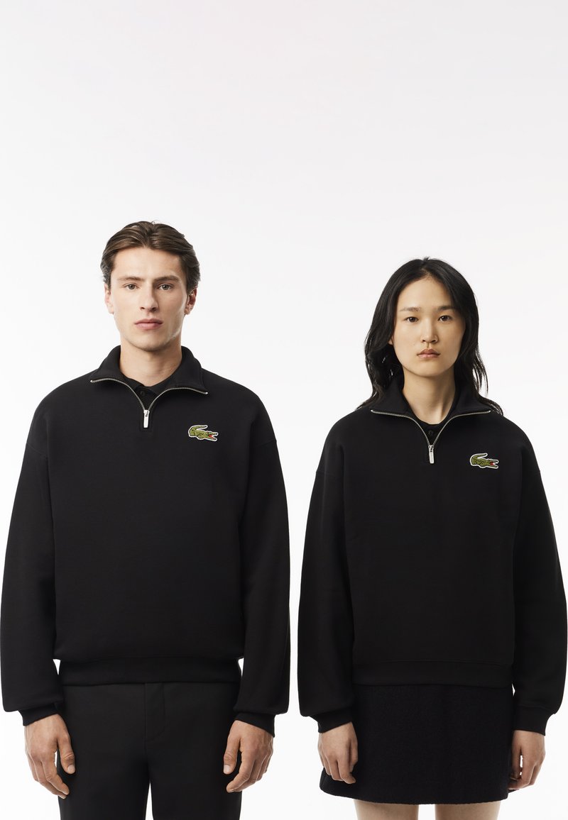 Sudaderas con cremallera negra de material suave, con un logo verde de cocodrilo de Lacoste en el lado izquierdo del pecho. Los cortes del modelo varían ligeramente.