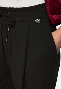 Black jogger pants with a drawstring waist, pleated front, side pockets, and a small branded label on the waistband. Smooth fabric texture.