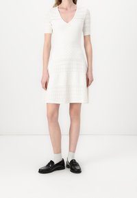 Robe blanche avec des manches courtes en dentelle et un décolleté en V, présentant des panneaux texturés sur toute la surface. Associée à des mocassins noirs et des chaussettes blanches.