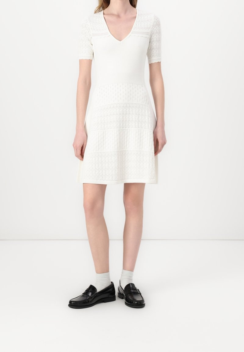 Robe blanche avec des manches courtes en dentelle et un décolleté en V, présentant des panneaux texturés sur toute la surface. Associée à des mocassins noirs et des chaussettes blanches.