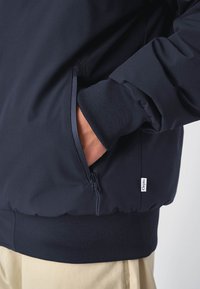 Marineblaue Steppjacke mit elastischen Bündchen und Saum, ausgestattet mit einer seitlichen Reißverschlusstasche und einem kleinen Logo-Tag auf der Vorderseite.