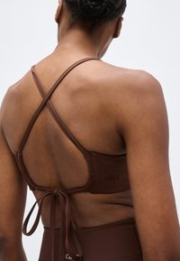 Γυναίκα που φοράει καφέ αθλητικό σουτιέν με σταυρωτούς ιμάντες και δεμένο πίσω, εμφανίζεται από τους ώμους μέχρι τη μέση σε απλό φόντο.