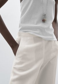 Bílý tílko spárované s volnými kalhotami v světle béžové barvě. Kalhoty mají boční kapsy, měkkou texturu a hladký povrch. Náhrdelník přidává detail.