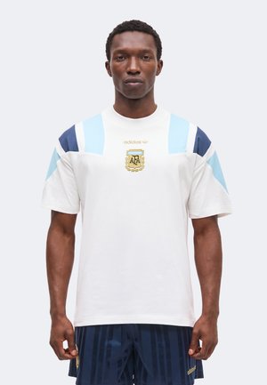 Mężczyzna w białej koszulce Adidas z jasnoniebieskimi i granatowymi panelami na ramionach oraz godłem Argentynskiego Związku Piłki Nożnej, w połączeniu z granatowymi krótkimi spodniami w paski.