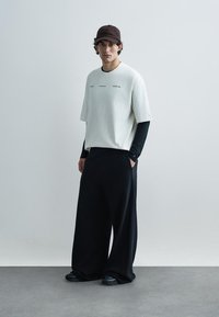 Gaiši pelēka t-krekla ar apaļu kaklu kārtojums virs melnas ar garām piedurknēm, kombinēta ar platām melnām biksēm un brūnu cepuri. Melni apavi noslēdz kopējo izskatu.