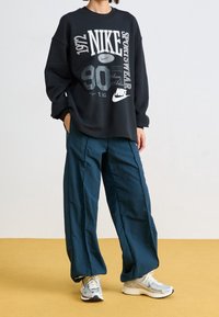 Černá mikina s výrazným bílým a šedým logem Nike, doplněná o volné tyrkysové kalhoty a světle šedé sportovní boty.