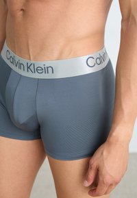 Boxer briefs grises hechos de tejido acanalado, con una cinturilla gris claro que presenta el logo "Calvin Klein". La vista en primer plano muestra el ajuste y la textura.