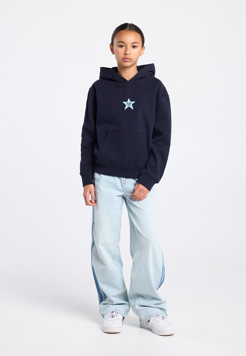 Marineblauwe hoodie met een lichtblauw sterrenlogo, gecombineerd met lichtblauwe wijde jeans met zijpanelen accenten. Witte sneakers.