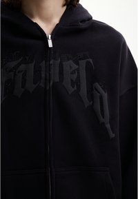 Haut à zip noir avec capuche, présentant un design de texte subtil en style gothique "SAVIEL" sur la poitrine, accompagné de petites clous décoratifs.