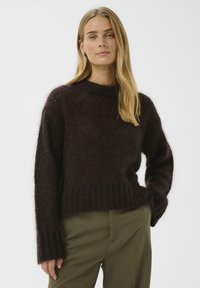 Brūns adīts džemperis ar rievotu apakšmalu un aprocēm, apaļu izgriezumu un teksturētu virsmu. Kombinēts ar olīvu zaļām biksēm.