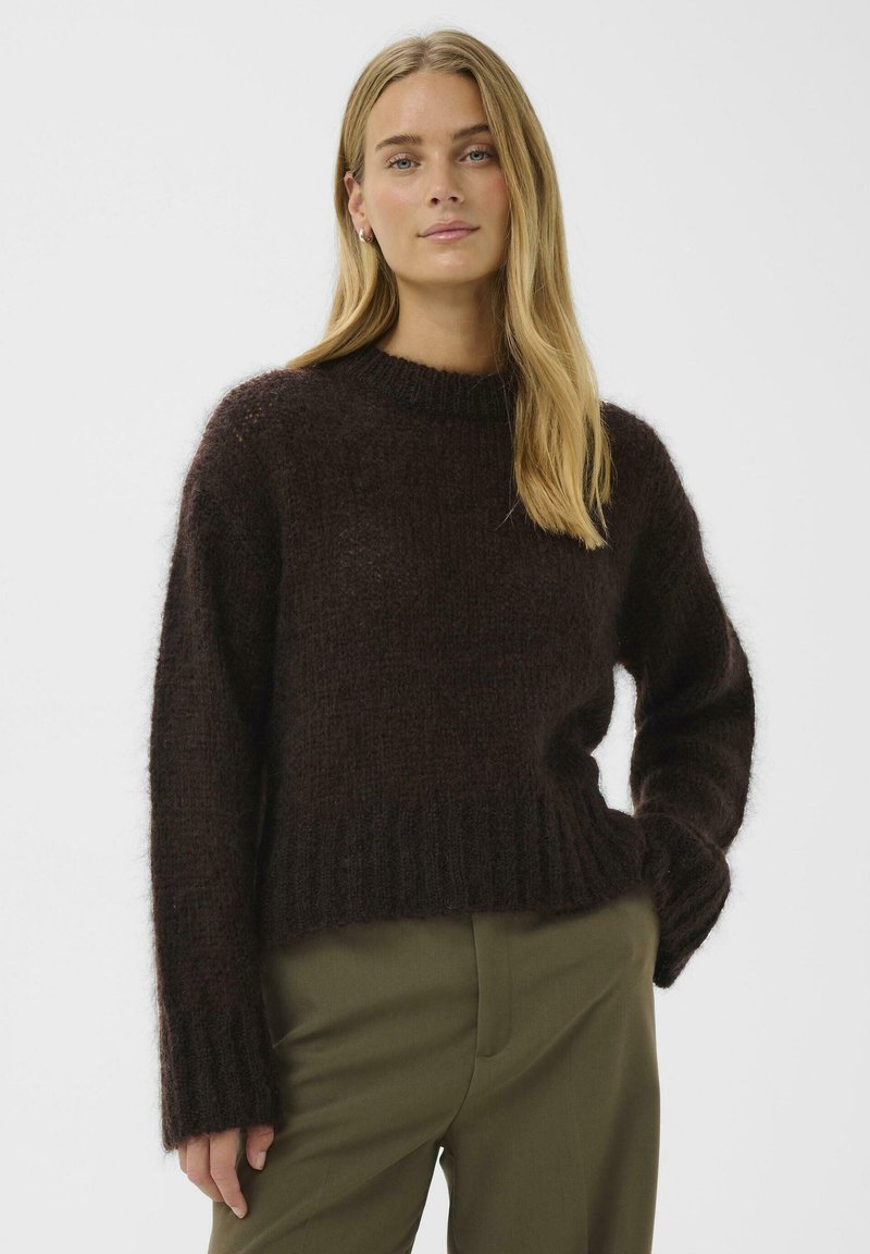 Brūns adīts džemperis ar rievotu apakšmalu un aprocēm, apaļu izgriezumu un teksturētu virsmu. Kombinēts ar olīvu zaļām biksēm.