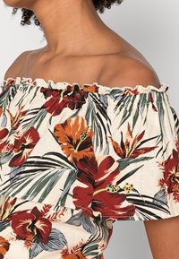 Femme portant un haut à épaules dénudées avec un imprimé floral tropical rouge, orange et vert sur fond crème.