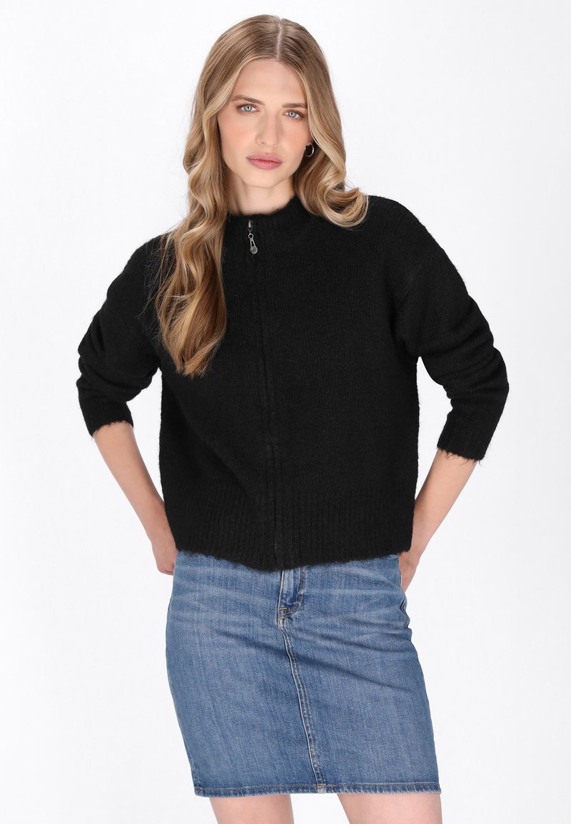 Maglione nero lavorato a maglia con colletto alto e zip frontale. Caratterizzato da maniche a tre quarti e orlo corto. Indossato sopra una gonna di jeans.