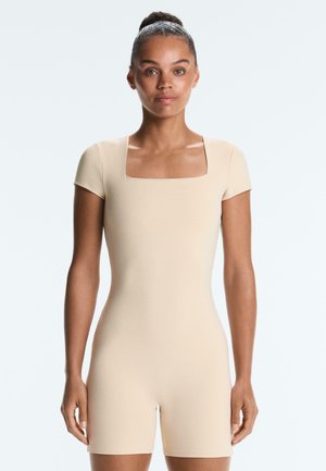 Kvinde med hår i knold iført en beige kortærmet, firkantet hals, tætsiddende romper, stående mod en ensfarvet lys baggrund.