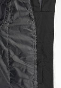 Primer plano del interior de una chaqueta negra que muestra un forro acolchado liso y un bolsillo de tela con cierre de botón a presión.
