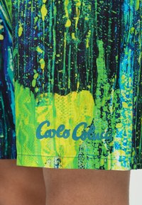 Bunte Badeshorts mit einem lebhaften Muster aus blauen, grünen und gelben Spritzern. Enthält das "Calo Coluca"-Logo in Hellblau.