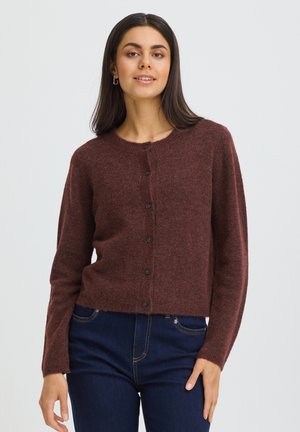 Jeune femme aux longs cheveux foncés portant un cardigan marron boutonné et un jean bleu foncé, debout devant un fond clair uni.