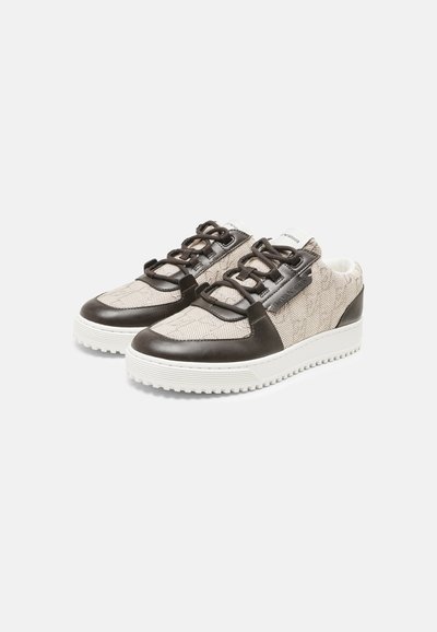 Paire de baskets basses avec tige en tissu beige, détails en cuir noir, lacets noirs et semelles épaisses blanches texturées sur un fond uni.