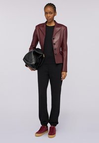 Vrouw in een burgundy leren blazer, zwarte top en broek, burgundy sneakers, met een grote zwarte leren tas tegen een effen achtergrond.