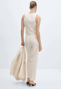 Haut sans manches en lin beige et pantalon assorti sur mesure. Le mannequin tient un blazer et porte des talons ouverts ; texture lisse et légère.