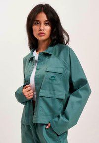 Grüne, taillierte Utility-Jacke mit großen Fronttaschen und Knopfverschluss. Glattes Material mit minimaler Textur, versehen mit einem Logo auf der linken Seite.