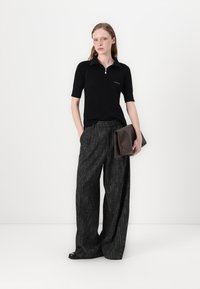 Polo negru cu dungi, cu mânecă scurtă și guler cu fermoar, purtat alături de pantaloni largi gri cu carouri. Modelul ține în mână o poșetă de mână maro.