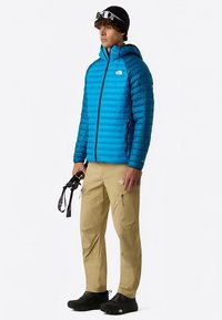 Chaqueta acolchada azul con rayas verticales, capucha y cremalleras. Pantalones beige con bolsillos con cremallera. Calzado negro y accesorio en la mano.