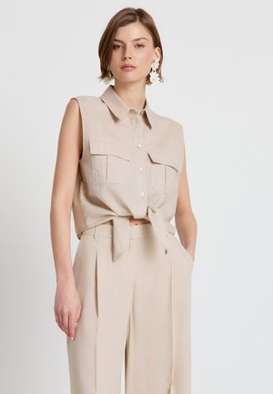 Femme portant un haut beige sans manches boutonné et noué à la taille, ainsi qu'un pantalon large taille haute assorti, accessoirisée avec de grandes boucles d'oreilles blanches en forme de fleurs.