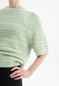 Mintgroene gebreide pullover met horizontale strepen en korte mouwen. Ribbelkraag en -zoom. Draag over zwarte broek, wat een ontspannen pasvorm laat zien.
