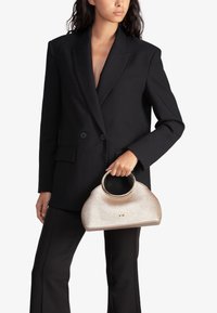 Bolso de mano metálico dorado con forma redondeada, que cuenta con un asa circular y una textura sutil. Se lleva con un blazer negro de doble botonadura y pantalones.