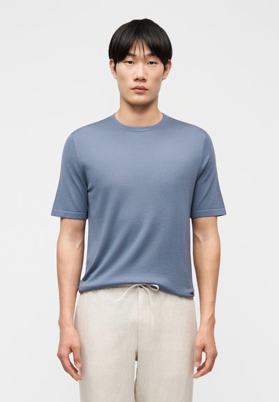 Jeune homme aux cheveux noirs raides portant une chemise bleu ajustée à manches courtes et un pantalon beige clair à cordon, debout devant un fond uni.