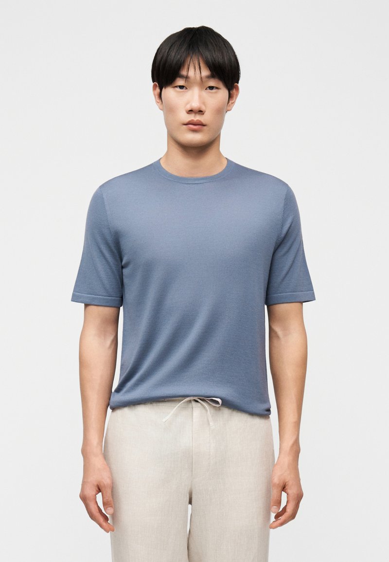 Νεαρός άνδρας με ίσια μαύρα μαλλιά, φοράει εφαρμοστό μπλε κοντομάνικο πουκάμισο και ανοιχτό μπεζ παντελόνι με κορδόνι, στέκεται μπροστά σε απλό φόντο.