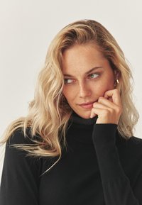 Schwarzer Rollkragenpullover mit gerippter Textur, getragen von einer Person mit welligem, blondem Haar. Weicher, glatter Stoff und figurbetontes Design.