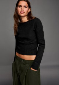 Fekete kötött pulóver bordázott szegéllyel és hosszú ujjakkal, lazán szabott olívazöld nadrággal, texturált felülettel párosítva.