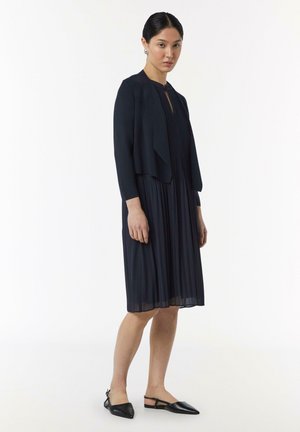 Abito a pieghe blu navy con scollatura rotonda e apertura frontale a chiave, abbinato a un cardigan lavorato a maglia cropped. Indossato con scarpe flat nere.