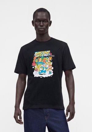 Hombre con una camiseta negra con un dibujo animado colorido y el texto "Don't take life too seriously" de pie frente a un fondo blanco.