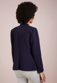 Personne aux cheveux bouclés vue de dos, portant un blazer bleu marine et un pantalon gris clair, sur un fond beige uni.