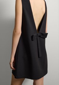 Vestido negro con un escote en V profundo y diseño sin mangas. Presenta un lazo en la cintura, hecho de una tela suave y ligeramente estructurada.