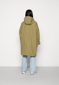 Długi płaszcz oliwkowozielony z kapturem, o prostym kroju i luźnym fasonie. Kurtka ma duże rękawy i jest zestawiona z jasnoniebieskimi jeansami.