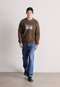 Brun sweatshirt med grafisk logotyp, grå detaljer och ribbade ärmslut, i kombination med ljusblå jeans och svarta skor. Modellen bär en svart mössa.